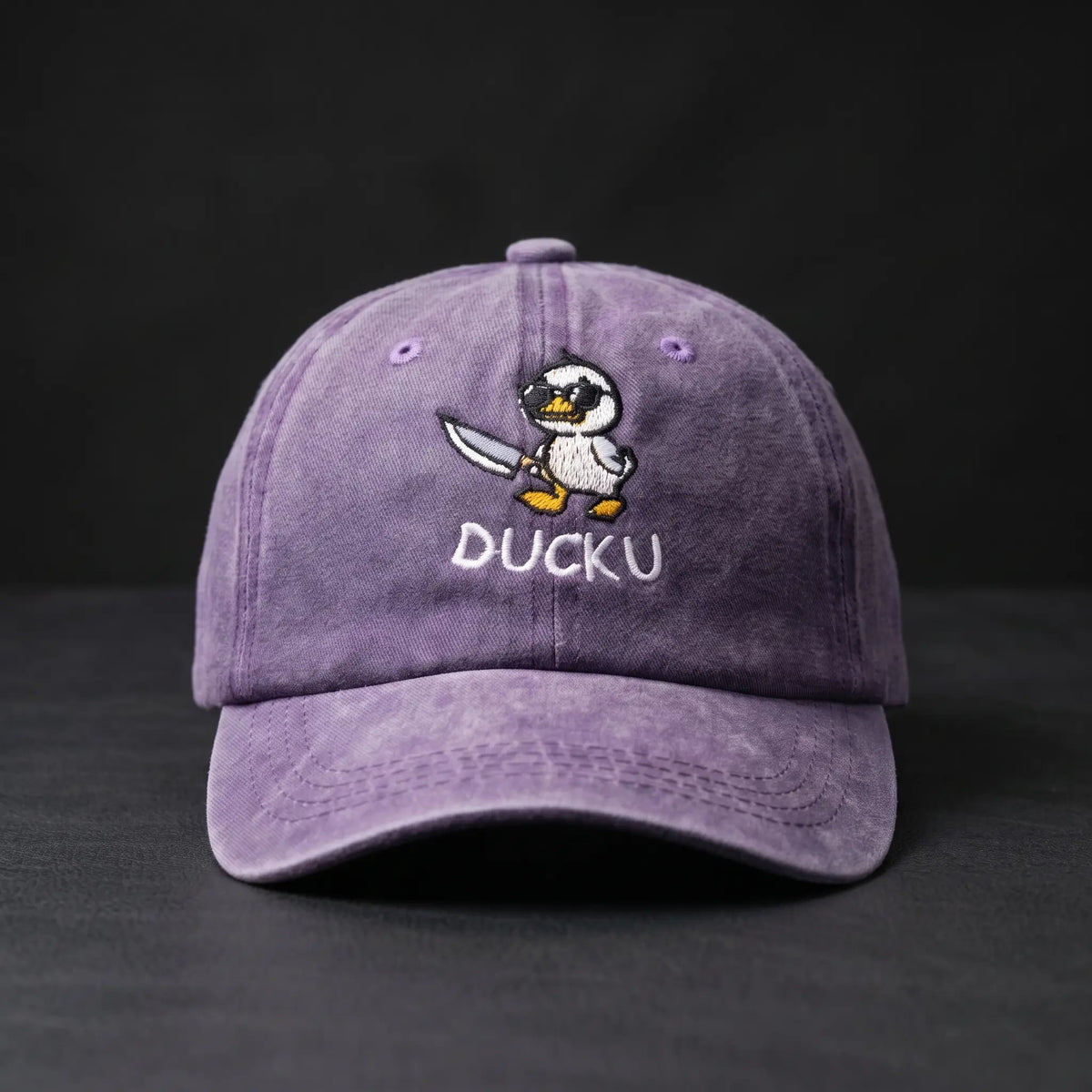 A purple washed cotton baseball cap featuring an embroidered cartoon duck character holding a knife with the text 'DUCKU' stitched below, set against a dark background.